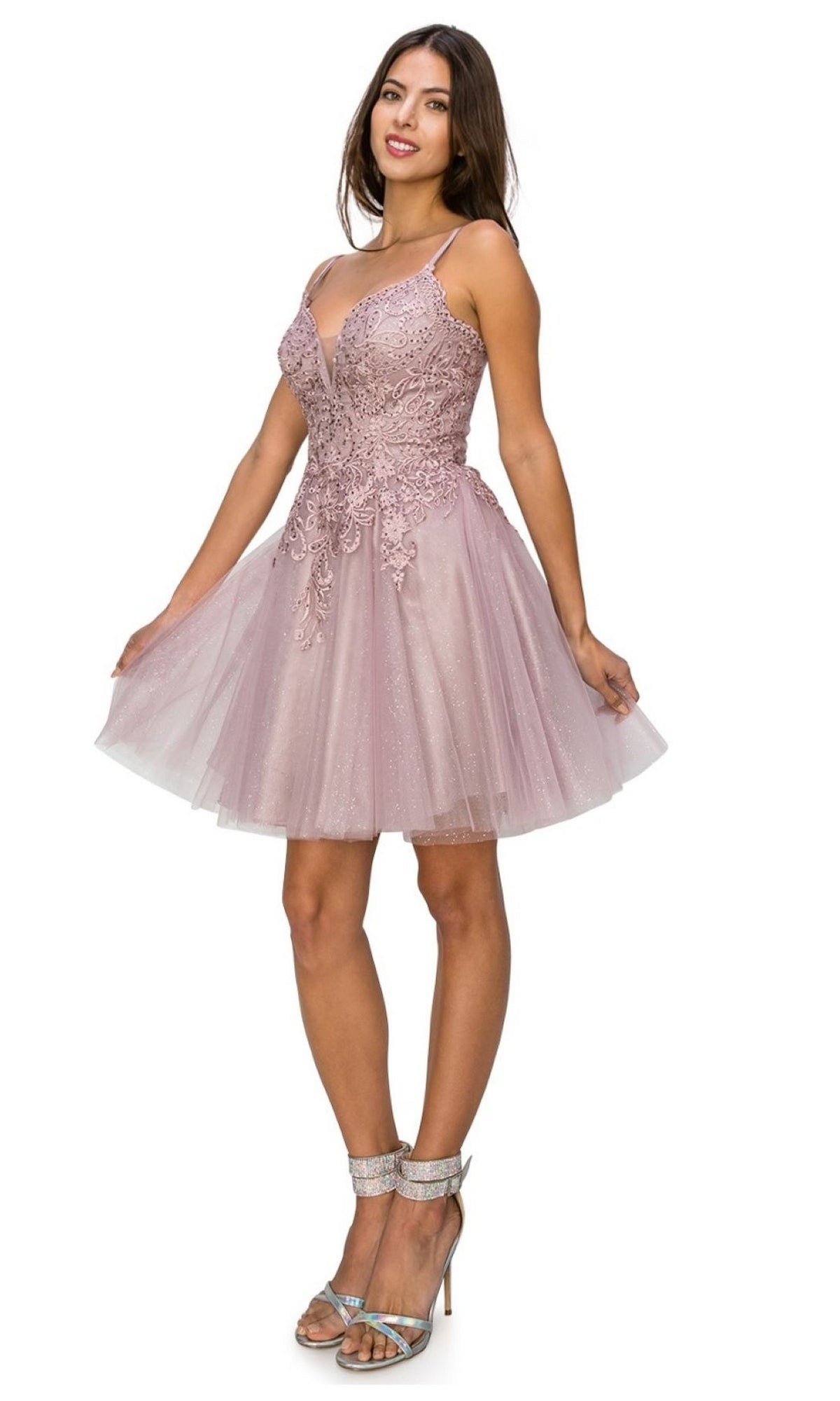 A woman wearing the Corset-Back Short Glitter Homecoming Dress 5125J, a sleeveless, blush pink, knee-length A-line party dress with lace detailing and a tulle skirt, paired with high-heeled sandals.