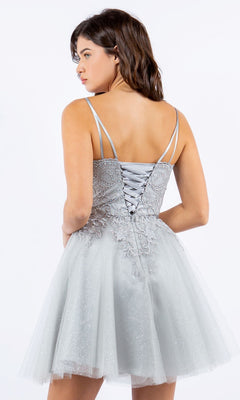 A woman is standing and facing away from the camera, wearing the Corset-Back Short Glitter Homecoming Dress 5125J in light gray with thin straps and a lace-up back.