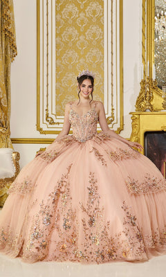 A young woman stands smiling at the camera in an ornate, gold-accented room, wearing the Quinceanera Dress 5131 by Dancing Queen in rose gold. The gown features a corset bodice and a flowing tulle ballgown skirt, with her tiara sparkling above the elaborately beaded design.