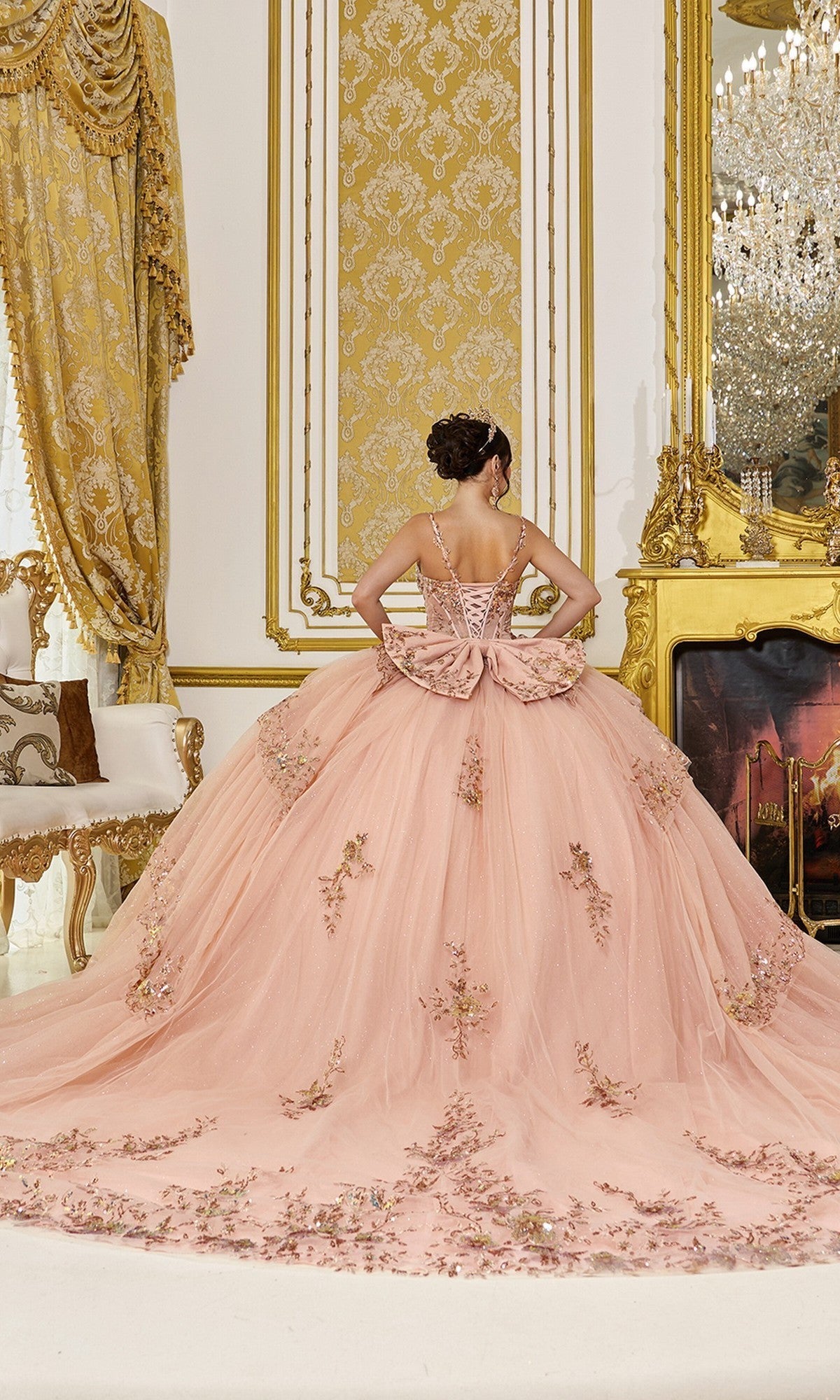 Wearing the Quinceanera Dress 5131 By Dancing Queen, a person stands facing a gold-accented fireplace in a lavishly decorated room, showcasing the gown’s ornate pink design, tulle ballgown skirt, and delicate floral embellishments.