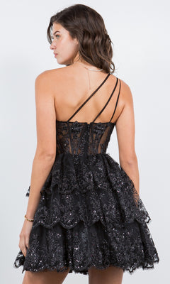 A woman faces away, wearing the One-Shoulder Short Tiered Homecoming Dress 5132J—a black, layered sequin party dress with lace accents and thin, crisscross straps—set against a plain light background.