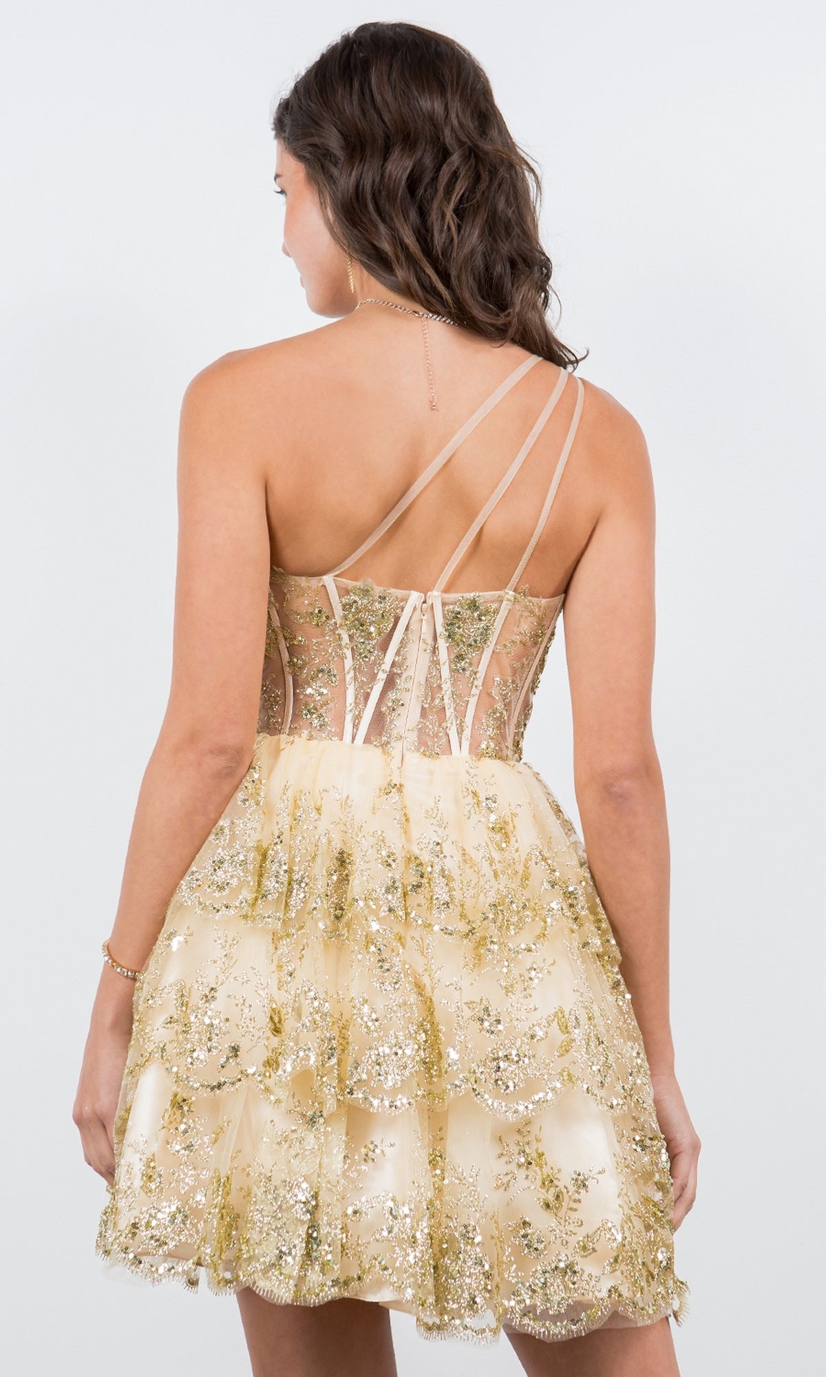 A woman wearing the One-Shoulder Short Tiered Homecoming Dress 5132J, featuring gold and beige sequins with a tiered skirt, stands facing away.