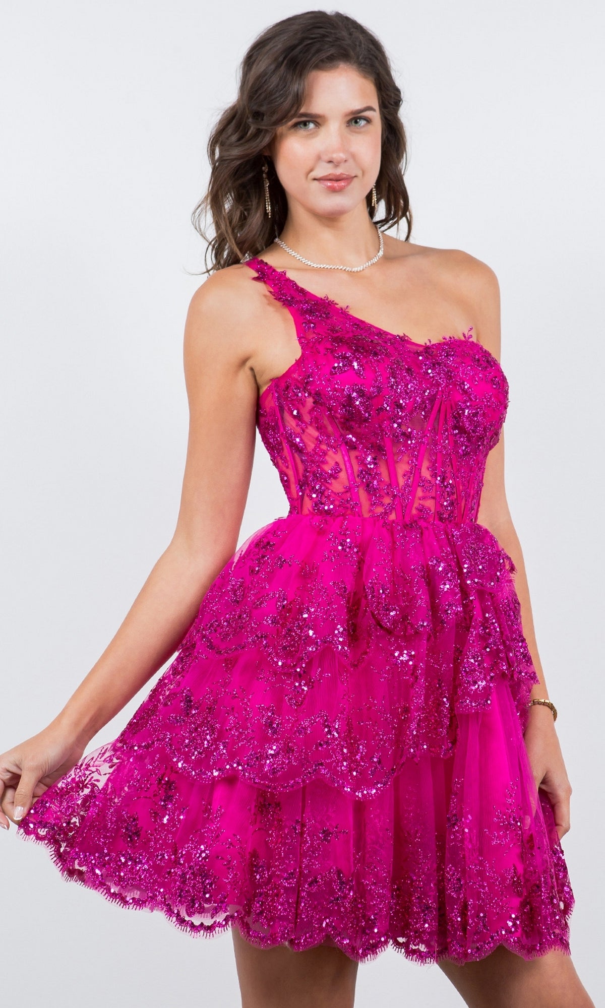 A woman poses against a plain background in the One-Shoulder Short Tiered Homecoming Dress 5132J, featuring bright pink fabric and layered sequined lace details.