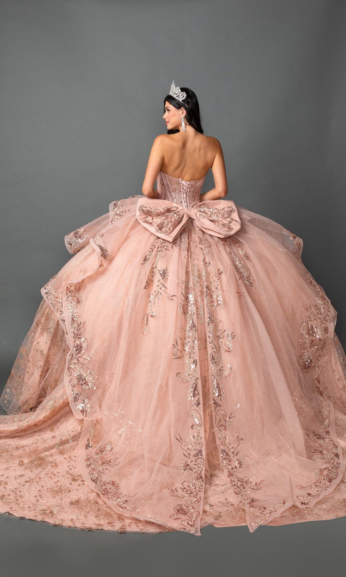 A woman stands against a plain gray background wearing the Quinceanera Dress 5133 By Dancing Queen—a pink, strapless ballgown with a beaded corset bodice, sparkling embroidery, and a large bow. She accessorizes her look with a tiara.