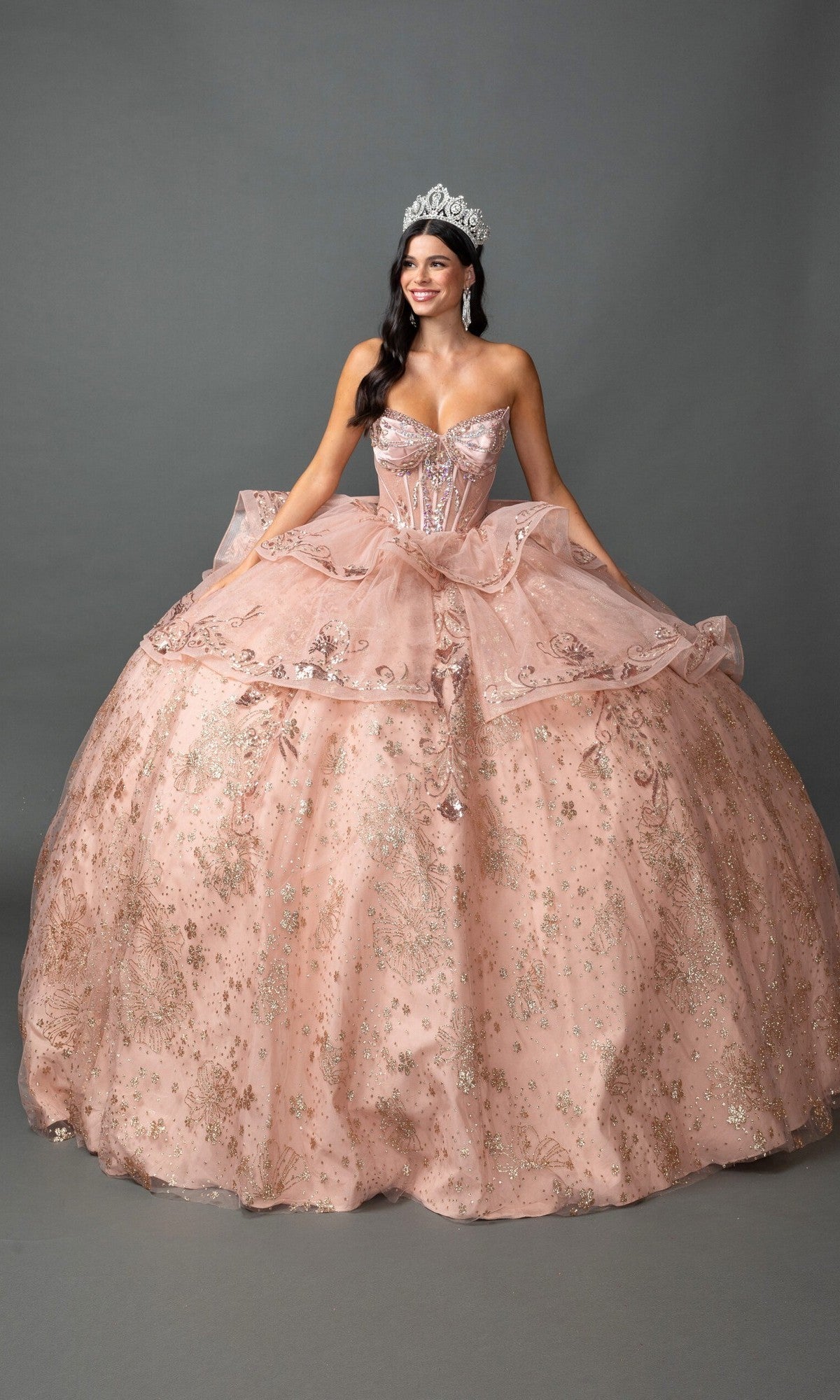 A woman wearing a silver tiara and the Quinceanera Dress 5133 by Dancing Queen in shimmering rose gold stands smiling against a plain gray background. The strapless ballgown features a beaded corset bodice, adding an elegant touch to her radiant look.