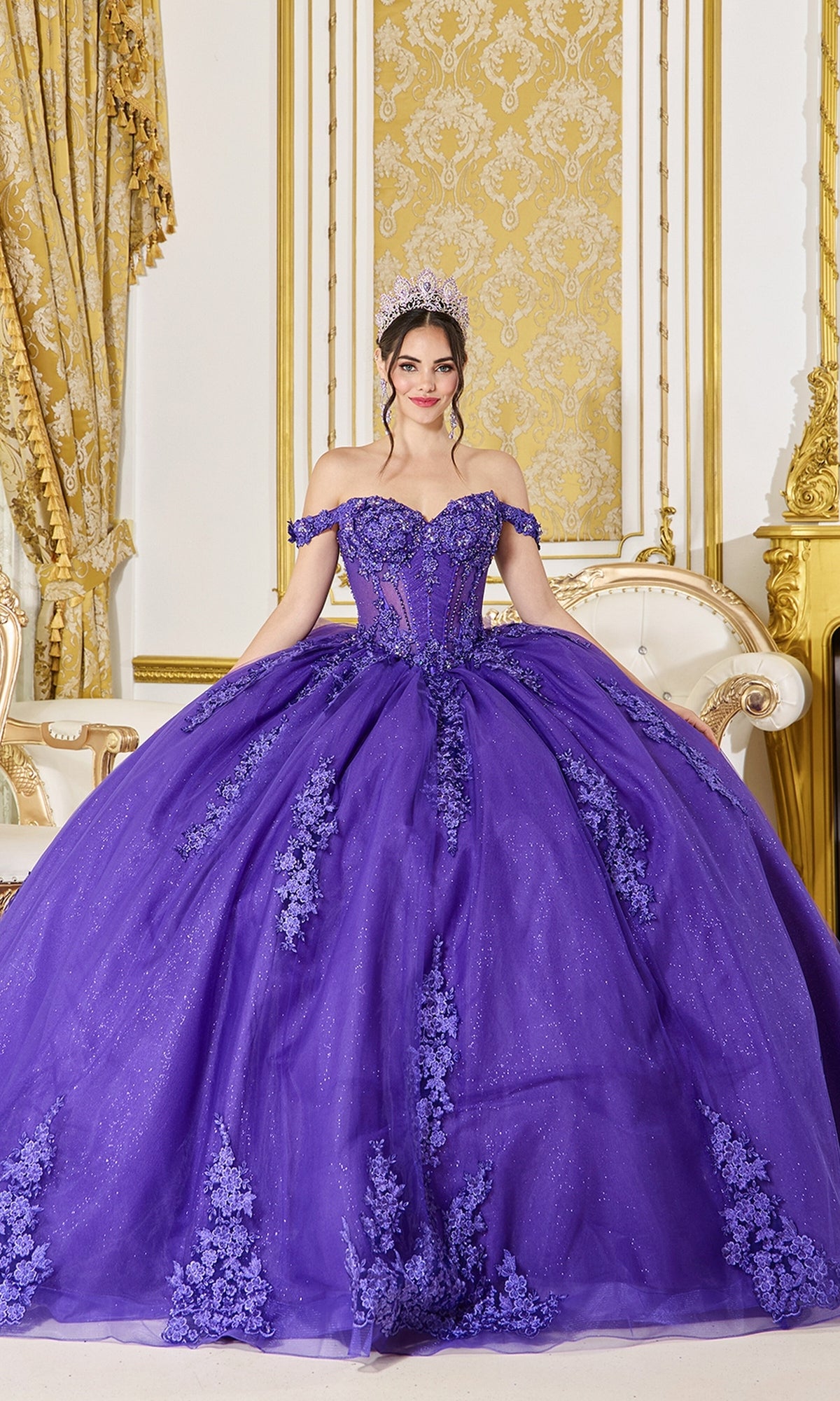 A woman poses indoors in a gold and white decorated room, wearing the Quinceanera Dress 5136 By Dancing Queen in purple, featuring floral lace details, a glitter tulle skirt, and a tiara.