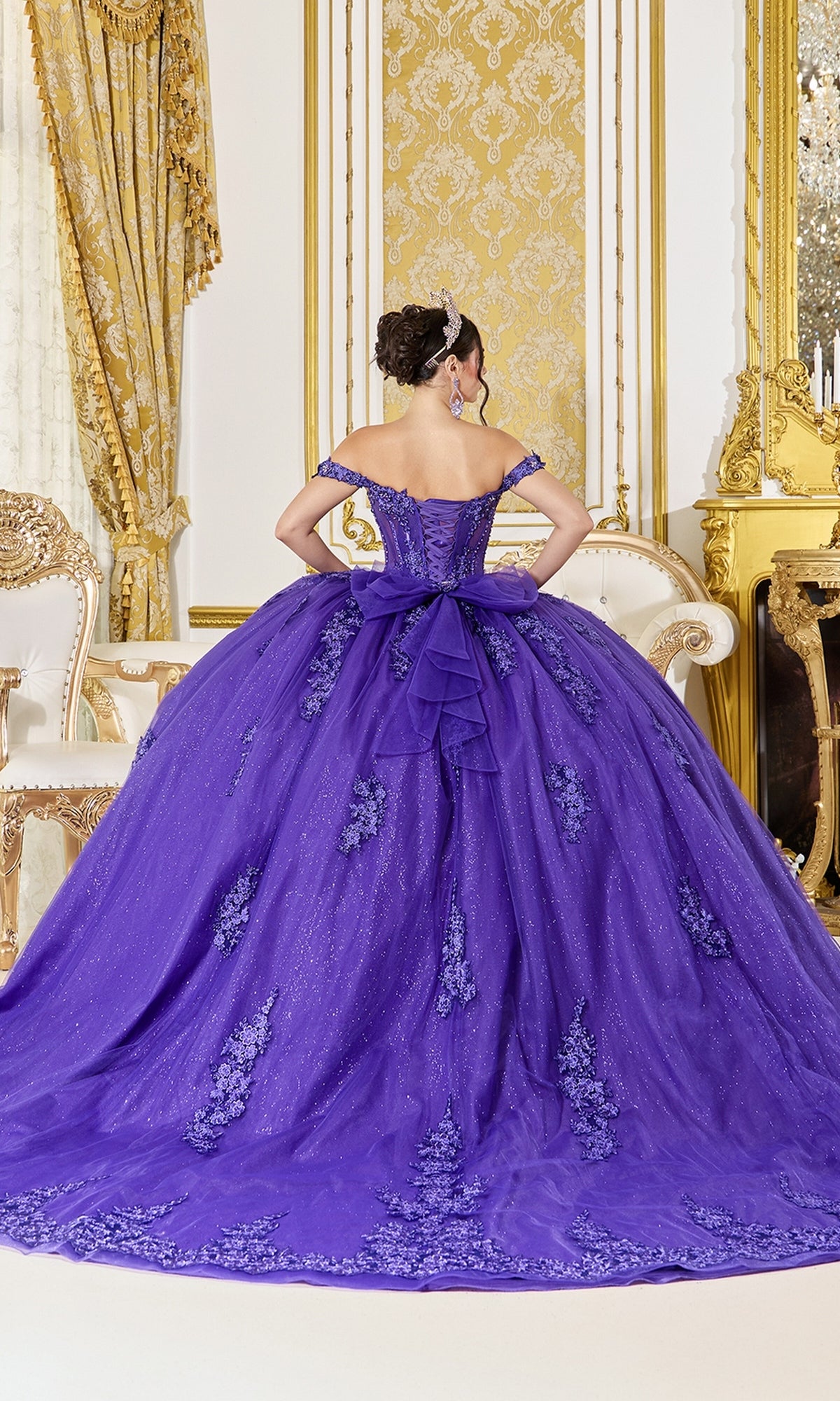 Wearing the Quinceanera Dress 5136 by Dancing Queen—a purple gown featuring lace details and a glitter tulle skirt—a woman stands facing away in an ornate room adorned with gold and white decor.