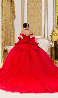 A woman wearing the Quinceanera Dress 5138 By Dancing Queen, an off-the-shoulder red ballgown, along with a tiara, sits on a white sofa seen from behind in a room adorned with ornate gold decor and patterned curtains.