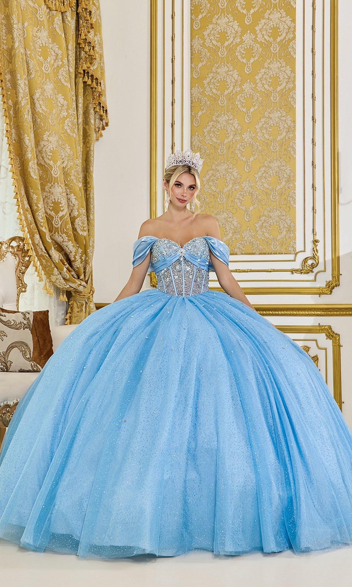 A woman stands in front of gold-patterned curtains and walls, wearing the Quinceanera Dress 5139 by Dancing Queen in sparkling Bahama Blue with an off-the-shoulder ball gown silhouette and a tiara.