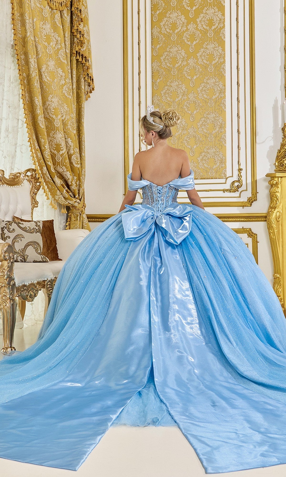 A person wearing the Quinceanera Dress 5139 By Dancing Queen, featuring ornate blue glitter and a large back bow, stands facing a gold and white decorated wall in an elegant room.