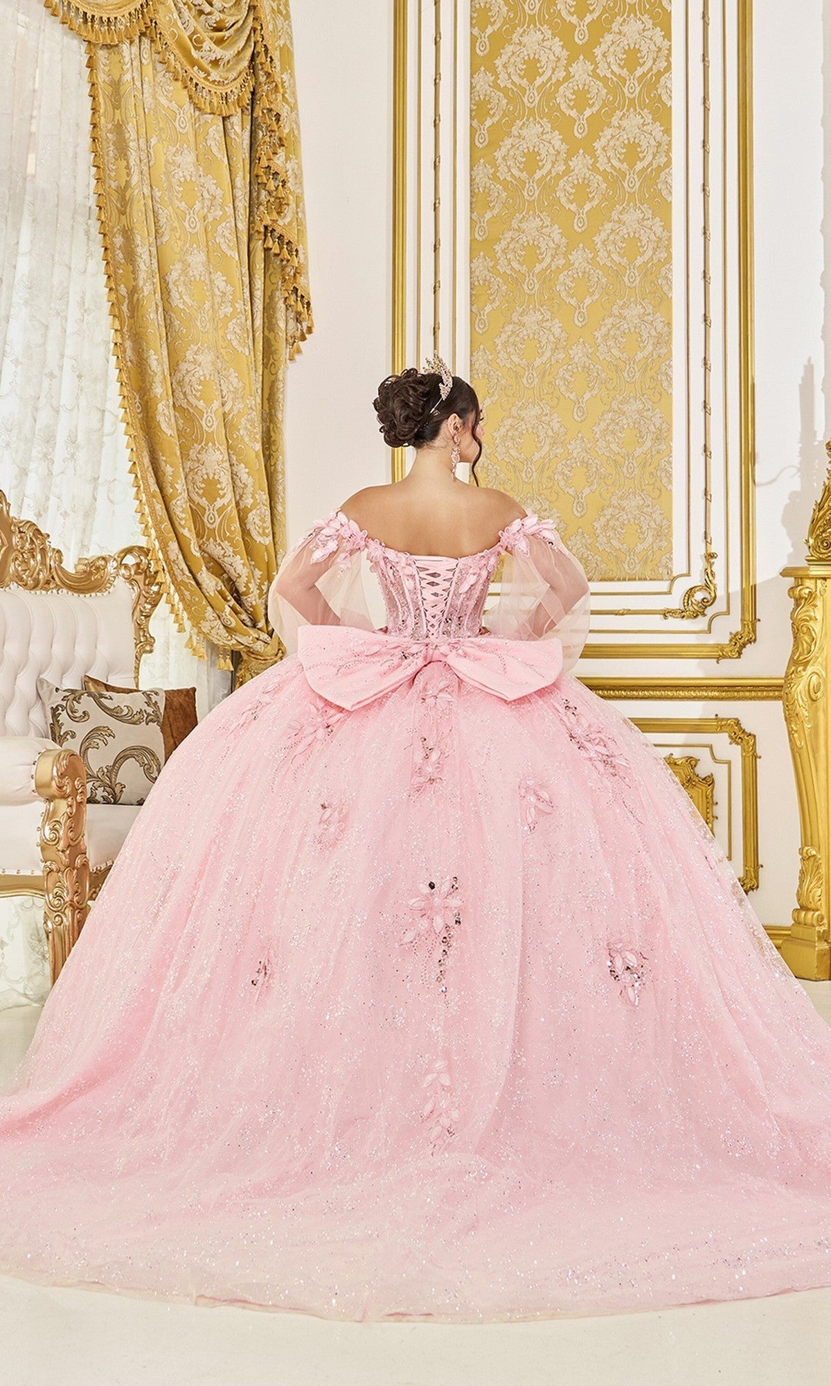 A woman stands facing away in an ornate, gold-accented room wearing the Dancing Queen Quinceanera Dress 5145, an off-the-shoulder pink ballgown adorned with delicate floral appliques and featuring a large bow on the back.