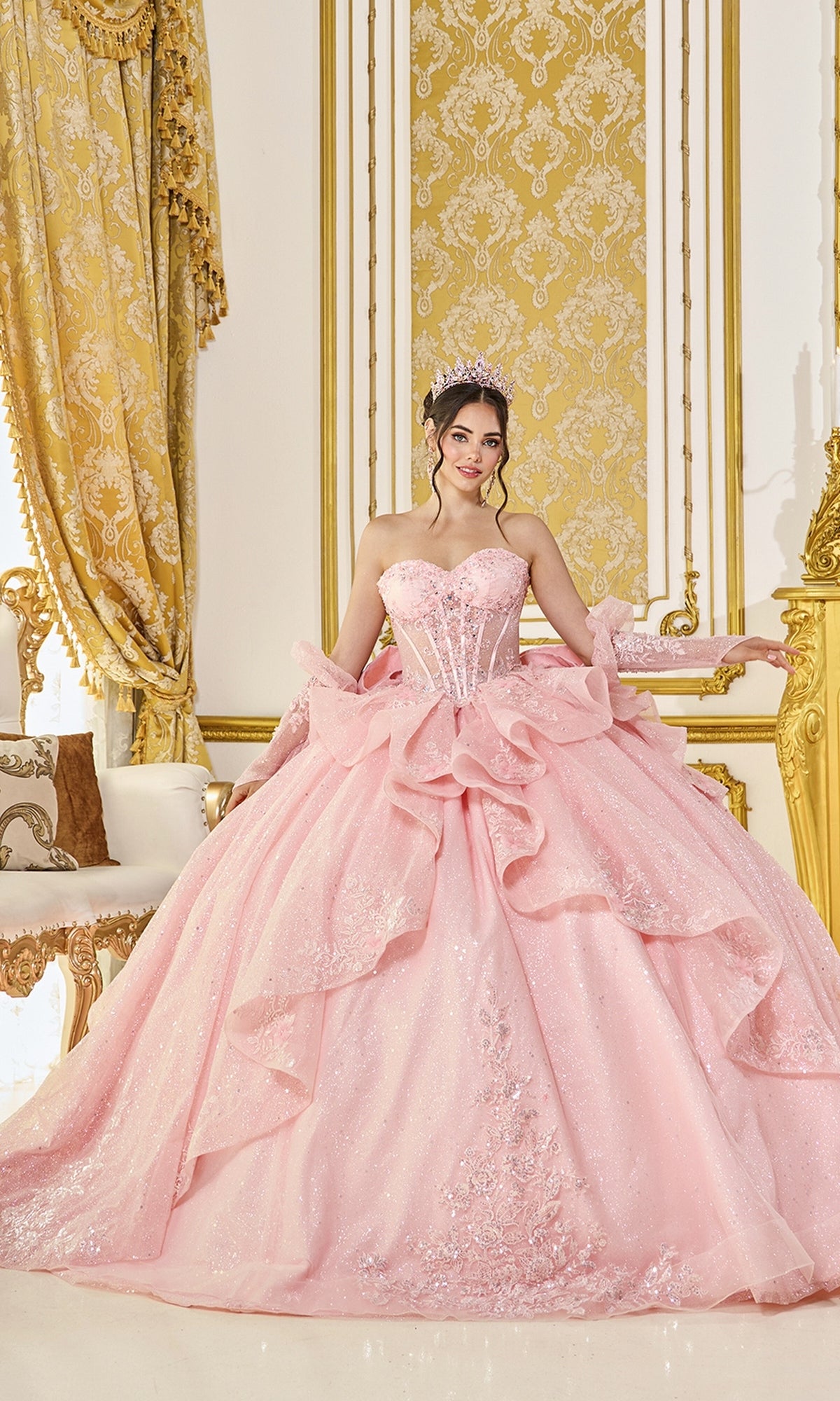 A young woman in the blush Quinceanera Dress 5152 by Dancing Queen, featuring a sparkling corset bodice and tiara, stands smiling at the camera in an ornate room adorned with gold and white decor.