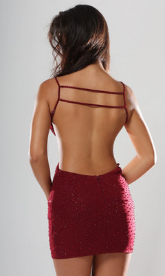 A woman faces away from the camera, showcasing the backless design of the Jovani 51553 short homecoming dress— a burgundy, spaghetti strap style featuring beaded embellishments and a deep v-neckline, set against a plain light background.