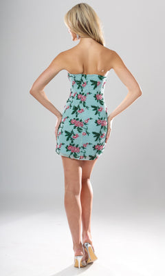 A woman stands facing away, wearing the Jovani 51557 short homecoming dress—a strapless bodycon mini dress featuring blue and pink floral patterns—paired with white high-heeled shoes against a plain background.