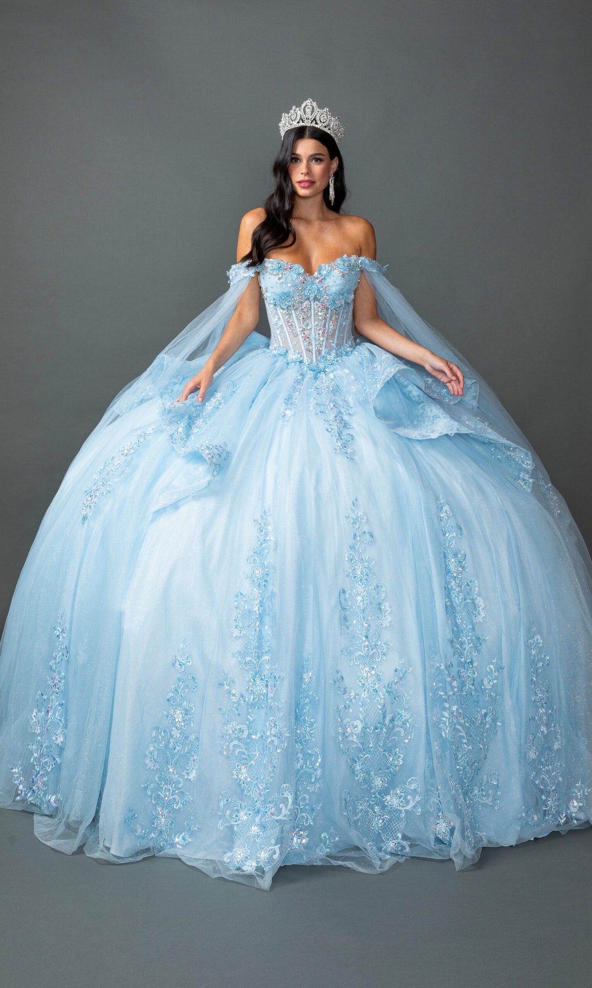 A woman wearing the Quinceanera Dress 5158 By Dancing Queen in Bahama Blue, featuring an off-the-shoulder design with floral details and an embroidered corset bodice, complemented by a tiara, stands against a plain gray background.