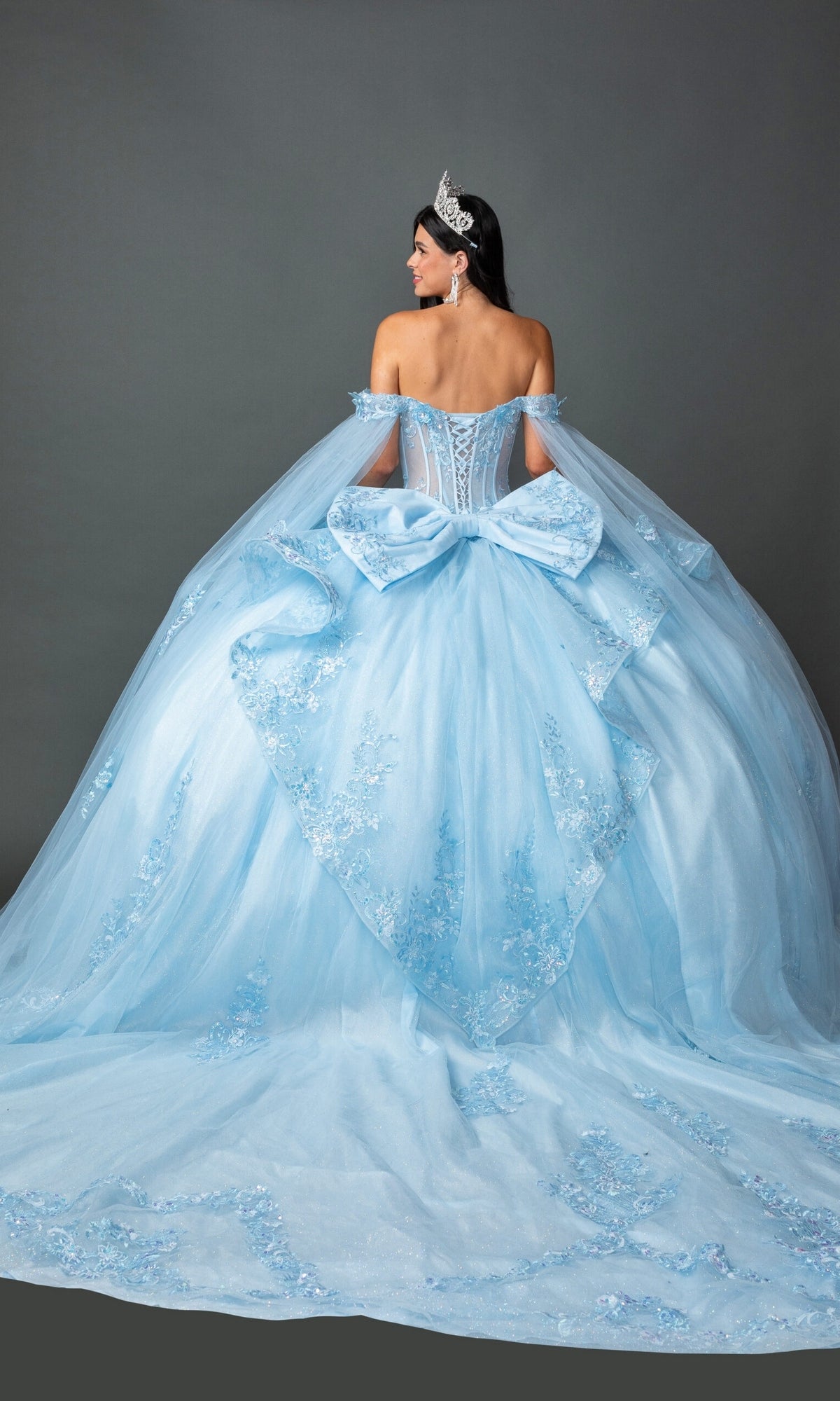 A woman stands facing away against a plain gray background, wearing the Quinceanera Dress 5158 by Dancing Queen—a light blue ball gown with off-the-shoulder sleeves, a large bow at the back, an elegantly embroidered corset bodice, and a flowing tulle skirt. She completes the look with a tiara.
