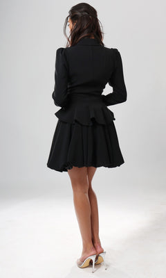 A woman stands on a white background, facing away from the camera, wearing the Short Homecoming Dress: Jovani 51602 in black—a fit-and-flare style featuring a ruffled hem—paired with beige high-heeled shoes.