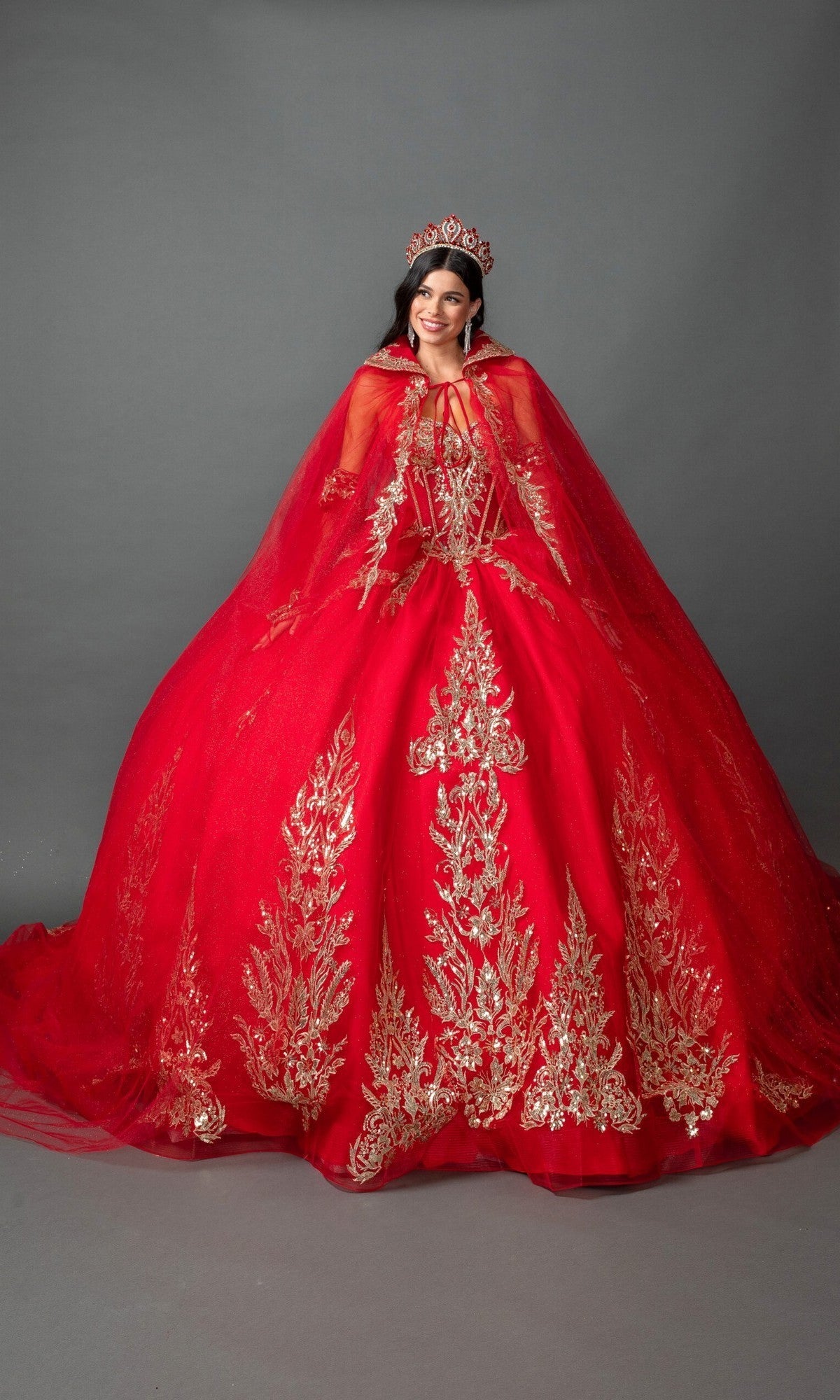 A woman is wearing the Quinceanera Dress 5161 by Dancing Queen—a stunning bright red strapless ballgown with gold embroidered appliques—paired with a matching red cape and a red tiara, standing against a plain gray background.