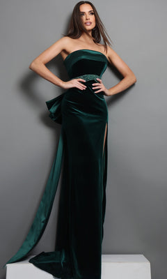 A woman wearing the Jovani 51648 long formal dress in hunter—a strapless dark green velvet gown with a side slit and a crystal-embellished waistband—stands against a gray background with one hand on her hip.