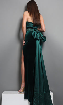 A woman stands on a white platform, facing away from the camera, wearing the Long Formal Dress: Jovani 51648—a strapless dark green velvet gown with a dramatic bow and train at the back. This exquisite dress is perfect for making an elegant statement at any formal event.