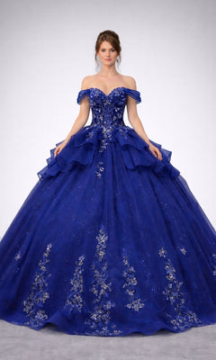 A woman wearing the Quinceanera Dress 5165 By Dancing Queen in navy, featuring an off-shoulder corset bodice adorned with floral embellishments and a full, layered skirt reminiscent of a glitter-tulle ballgown, stands against a plain background.