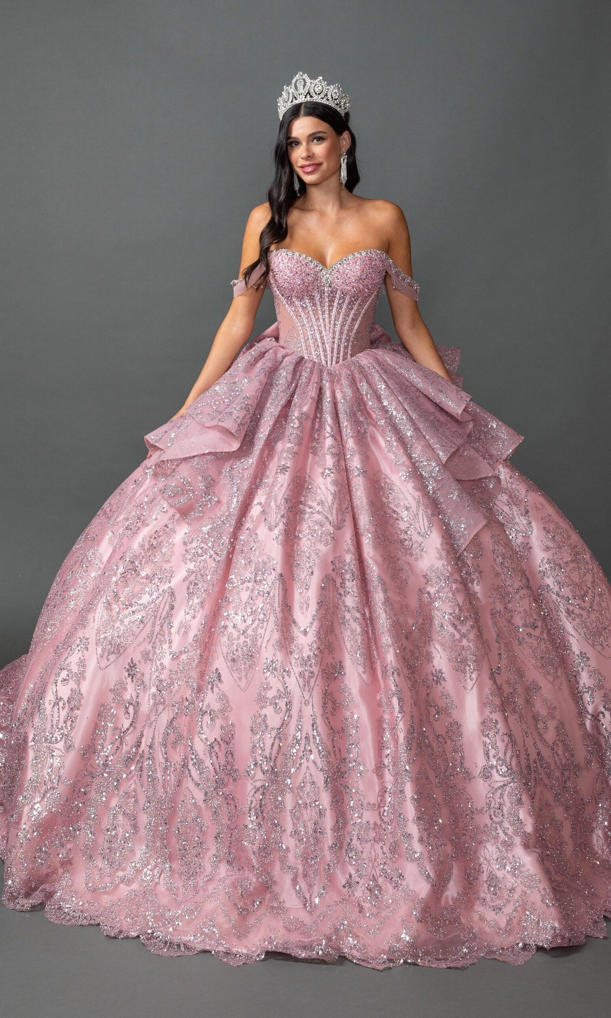 A woman wearing the Quinceanera Dress 5169 by Dancing Queen in Dusty Pink—with a sparkling beaded corset bodice and ballgown skirt—stands against a gray background, holding the sides of her dress and smiling at the camera.