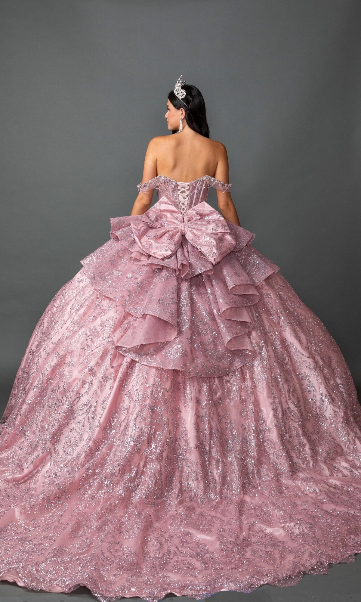 A woman, seen from behind against a gray background, wears the Quinceanera Dress 5169 By Dancing Queen—an off-shoulder, sparkling pink ballgown featuring a full skirt, oversized bow, and tiara.