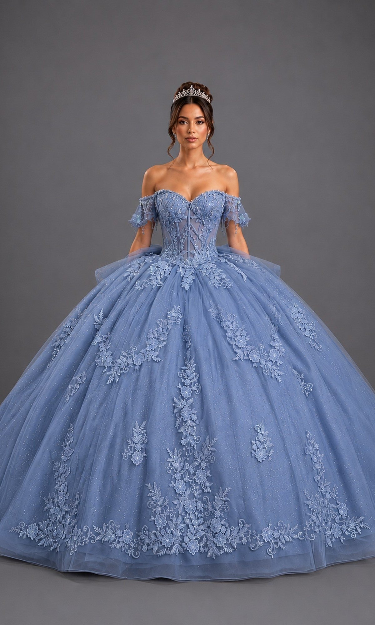 A woman wearing the Quinceanera Dress 5171 By Dancing Queen in dusty blue, featuring off-shoulder floral lace detailing and a tiara, stands against a plain gray background.