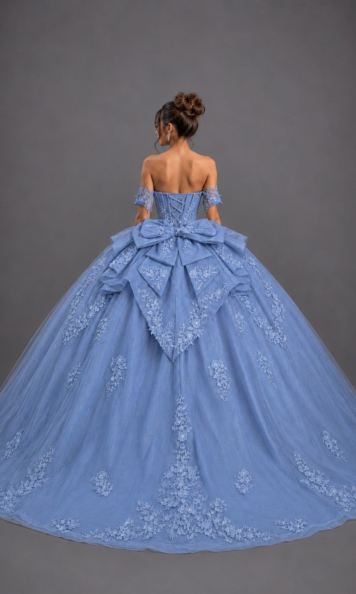 A woman stands against a plain gray background, wearing the Quinceanera Dress 5171 by Dancing Queen—a dazzling blue off-the-shoulder ballgown adorned with elegant floral embroidery, a large bow at the back, and glitter tulle that lends sparkling glamour to the classic silhouette.
