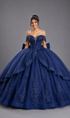 A woman stands smiling in the Quinceanera Dress 5172 by Dancing Queen—a navy, off-shoulder ball gown adorned with lace and sequin details and featuring a corset-style bodice—against a plain gray backdrop.