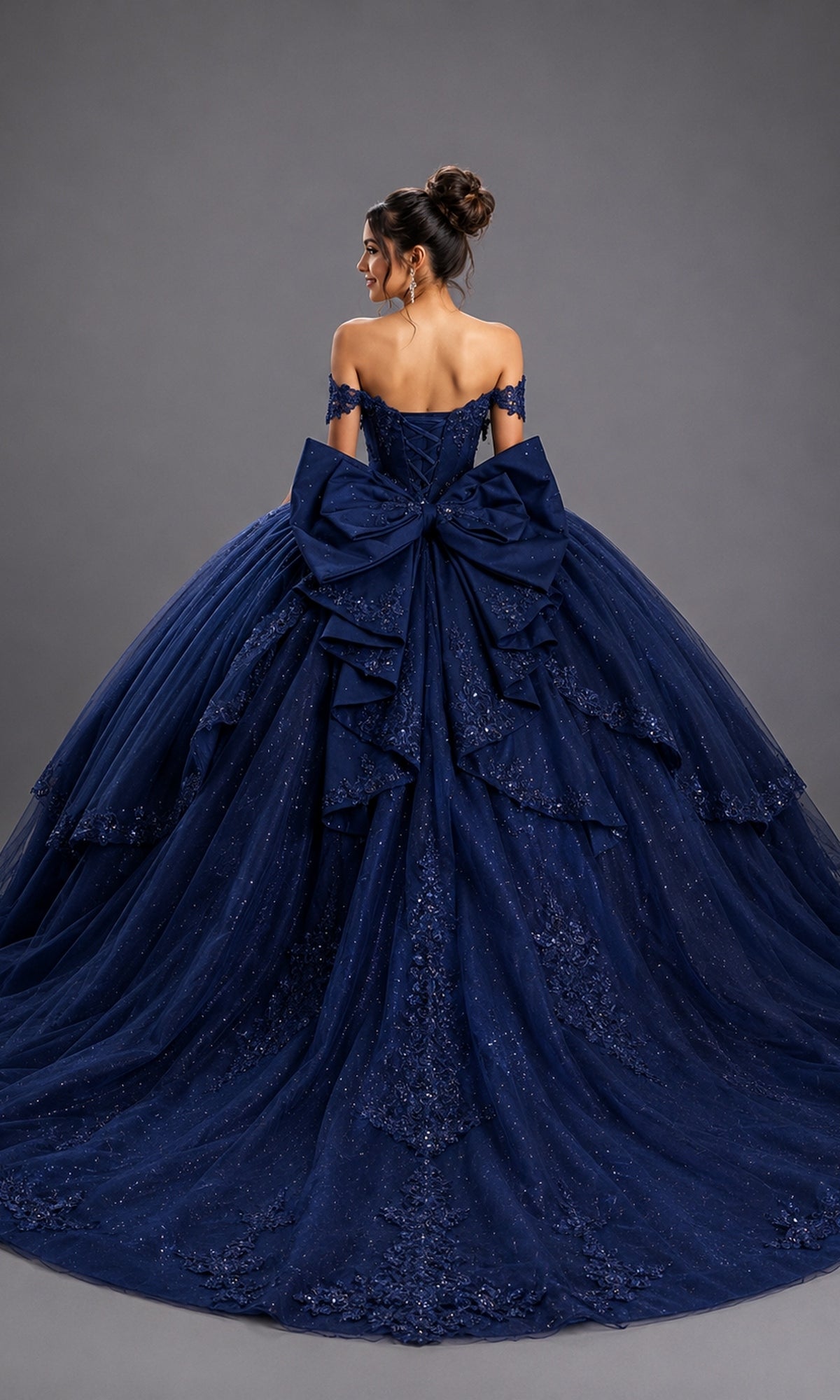 A woman poses with her back to the camera against a plain gray background, wearing the Quinceanera Dress 5172 by Dancing Queen—an off-the-shoulder ballgown crafted from deep blue glitter tulle, featuring a corset-style bodice, intricate detailing, and a large decorative bow.