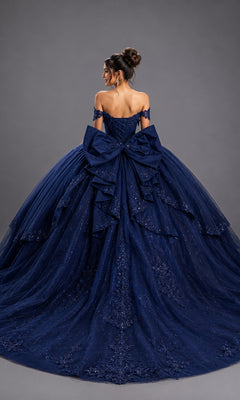 A woman poses with her back to the camera against a plain gray background, wearing the Quinceanera Dress 5172 by Dancing Queen—an off-the-shoulder ballgown crafted from deep blue glitter tulle, featuring a corset-style bodice, intricate detailing, and a large decorative bow.