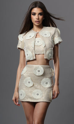 A woman wears the Jovani 51734 Short Homecoming Dress in cream, featuring textured embellishments and large white floral designs, as she stands against a plain gray background.