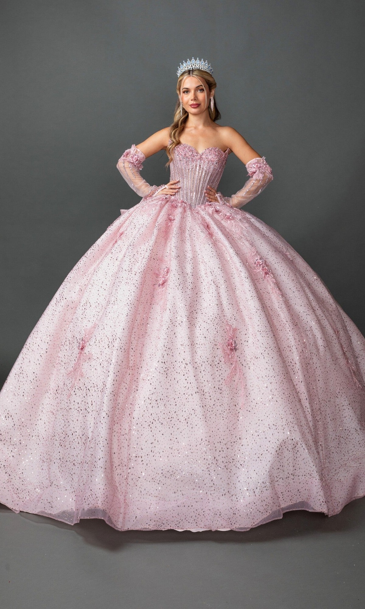 A woman stands with hands on hips, wearing the Quinceanera Dress 5173 by Dancing Queen in a sparkling blush hue, featuring a beaded corset bodice and long gloves, topped off with a tiara, against a plain gray background.