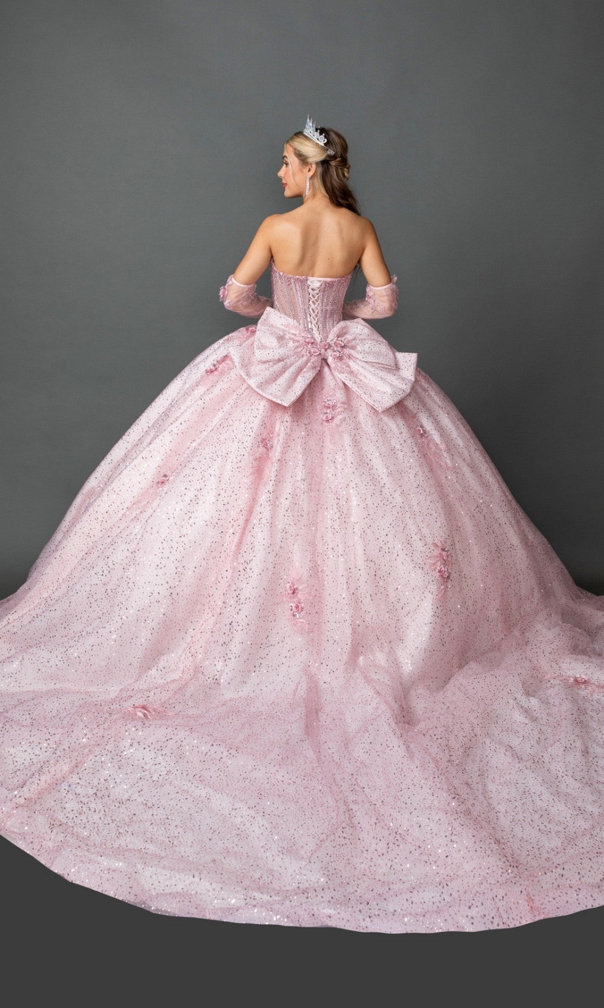 A woman stands facing away against a plain gray background, wearing the Quinceanera Dress 5173 By Dancing Queen—a sparkling pastel pink gown with a beaded corset bodice, large bow accent on the back, tiara, and long matching gloves.