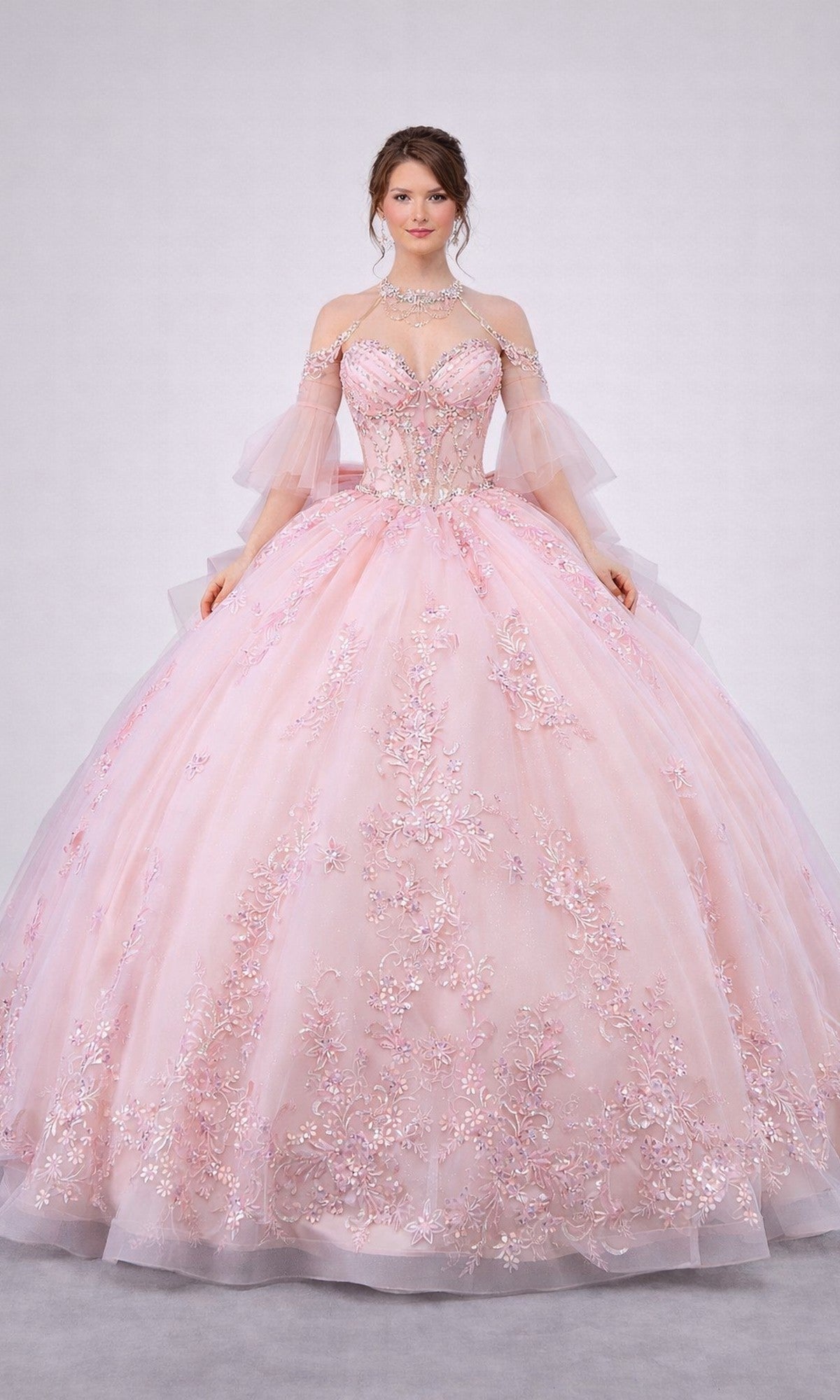 A woman stands in the Quinceanera Dress 5179 By Dancing Queen, a blush gown featuring intricate embroidery, off-the-shoulder tulle sleeves, and a full ballgown tulle skirt, against a plain light gray background.