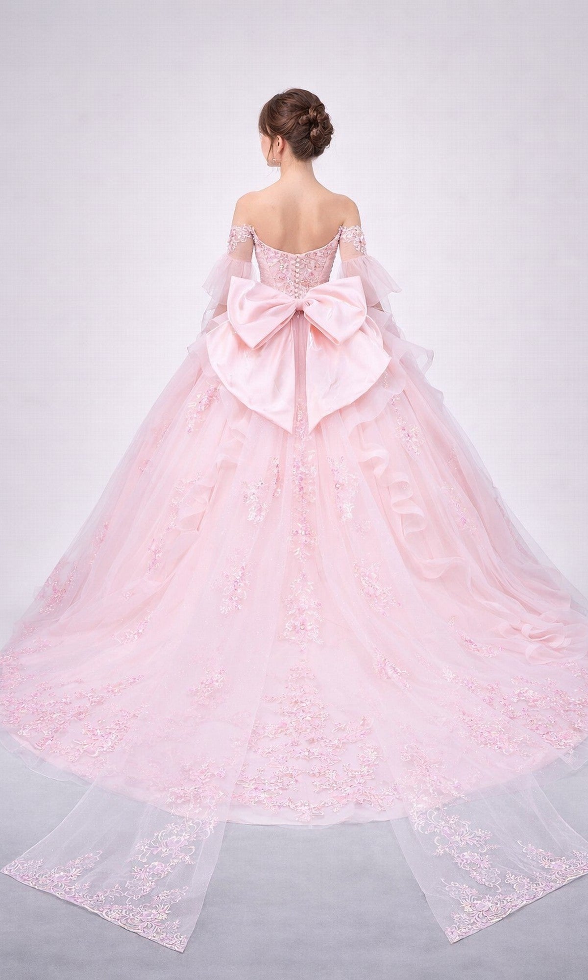 A woman is seen from behind wearing the Quinceanera Dress 5179 by Dancing Queen: an off-the-shoulder, floor-length pink gown adorned with intricate floral embroidery, featuring a dramatic tulle skirt, a structured corset bodice, and a large bow at the back. The dress’s long, flowing train trails gracefully as she stands turned away.
