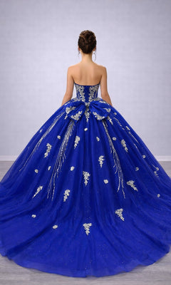 A woman stands facing away against a plain background, wearing the Quinceanera Dress 5183 by Dancing Queen—a blue strapless gown featuring silver floral embellishments and a structured corset bodice.