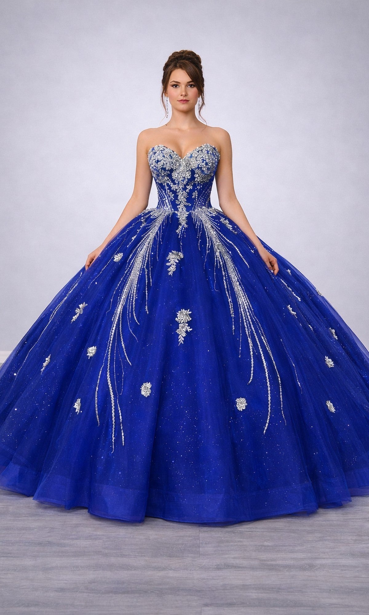A woman wearing the Quinceanera Dress 5183 By Dancing Queen in royal blue, featuring a strapless design with silver embellishments and a glitter tulle ballgown skirt, stands against a plain, light gray background.