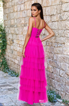 A woman stands outside on a stone path by a brick wall, wearing the Blondie Nites 5187BN long hot pink prom dress with thin straps, ruched sweetheart bodice, tiered tulle skirt, and lace-up back—a gorgeous under-$200 choice.