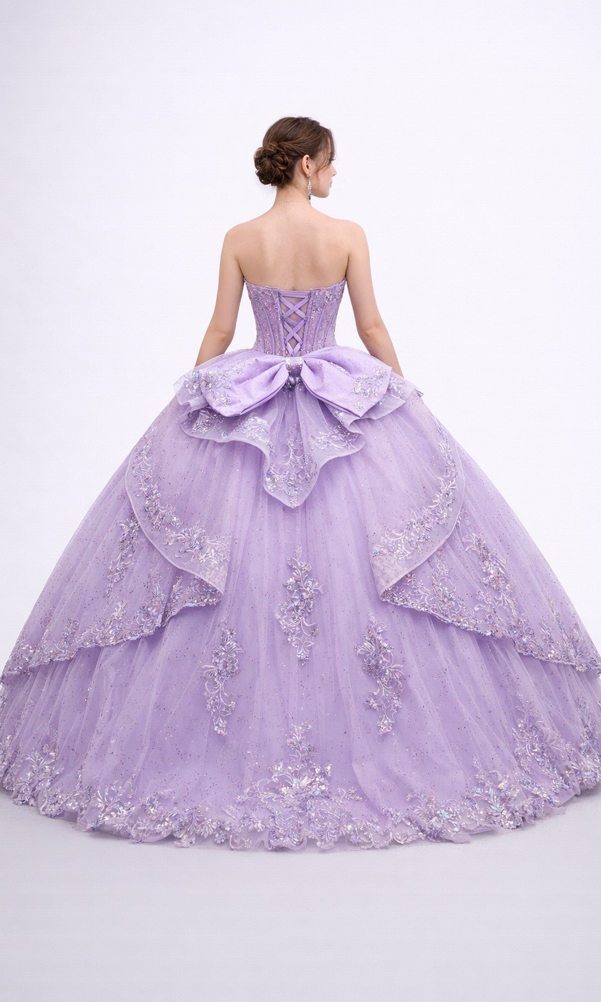 A woman is seen from the back against a plain white background wearing the Quinceanera Dress 5188 by Dancing Queen, which features a strapless corset bodice adorned with lace and beading, a large bow accent, and a voluminous ballgown skirt.