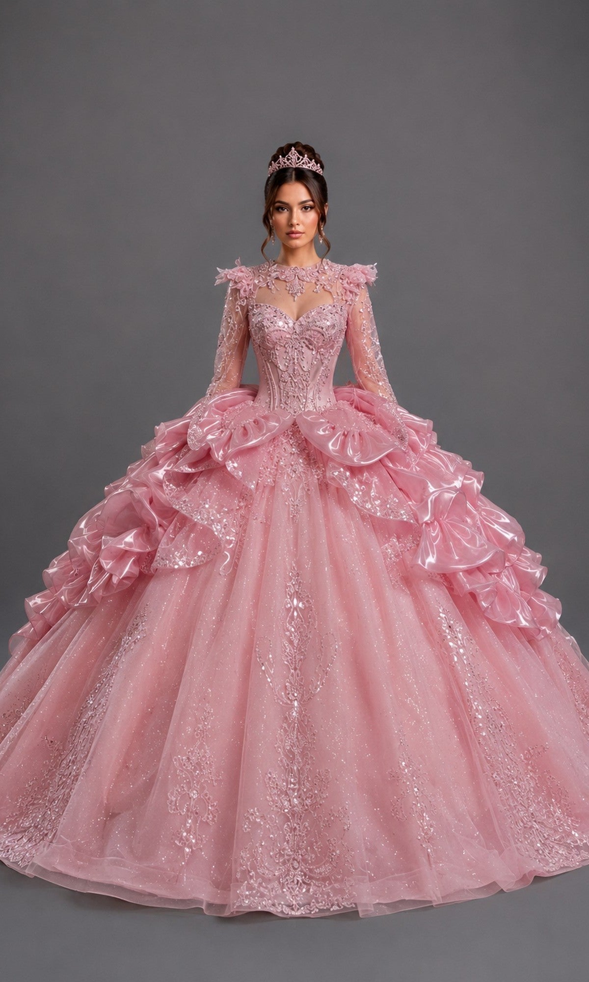 A woman stands against a plain gray background wearing the Quinceanera Dress 5189 By Dancing Queen in pink, featuring a voluminous silhouette with lace details, an embellished fitted bodice, long sheer sleeves, and a matching tiara.