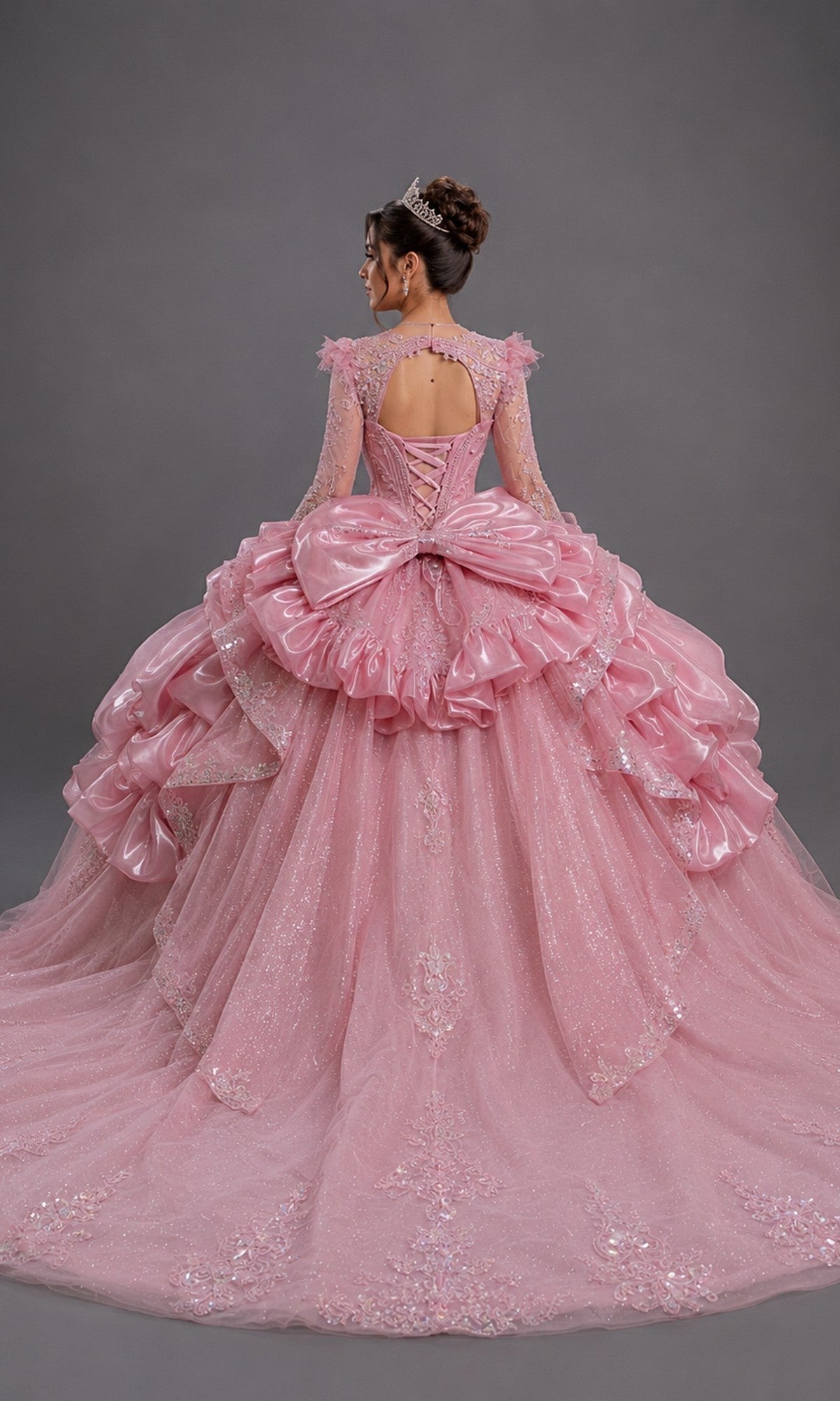 A woman faces away, wearing the Quinceanera Dress 5189 by Dancing Queen—a sparkling pink gown adorned with ruffles, a large bow, and lace details, paired with a tiara. The fitted bodice is beautifully embellished and the dress showcases layers of glitter tulle. The background is plain gray.