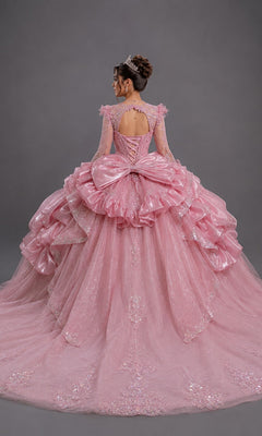 A woman faces away, wearing the Quinceanera Dress 5189 by Dancing Queen—a sparkling pink gown adorned with ruffles, a large bow, and lace details, paired with a tiara. The fitted bodice is beautifully embellished and the dress showcases layers of glitter tulle. The background is plain gray.