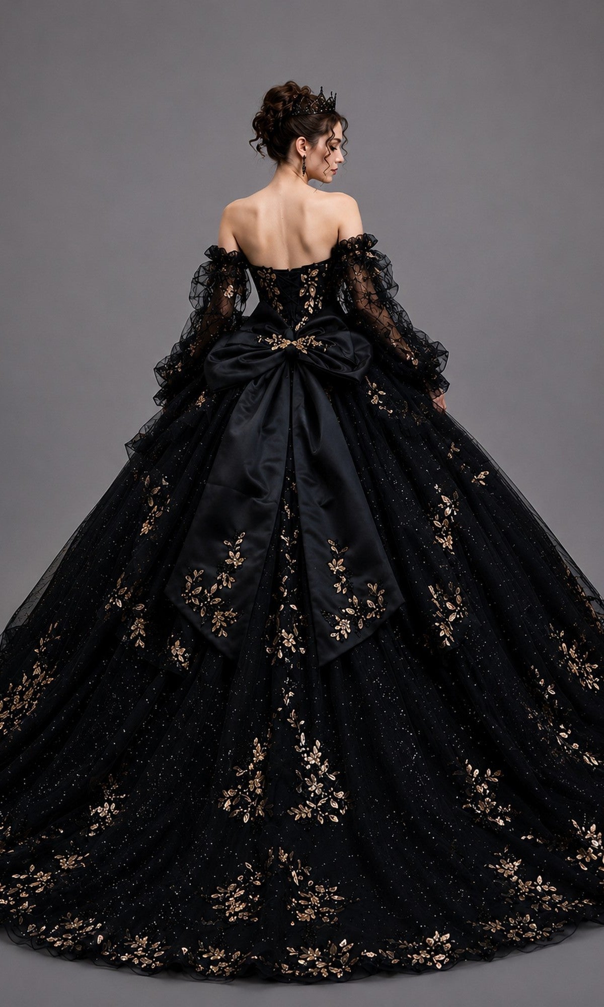 A person wearing the Quinceanera Dress 5190 By Dancing Queen—an off-shoulder black ballgown adorned with intricate gold leaf appliques, featuring a large bow on the back and paired with a tiara—stands against a plain gray background, creating a stunning and unforgettable look for any Quinceanera celebration.