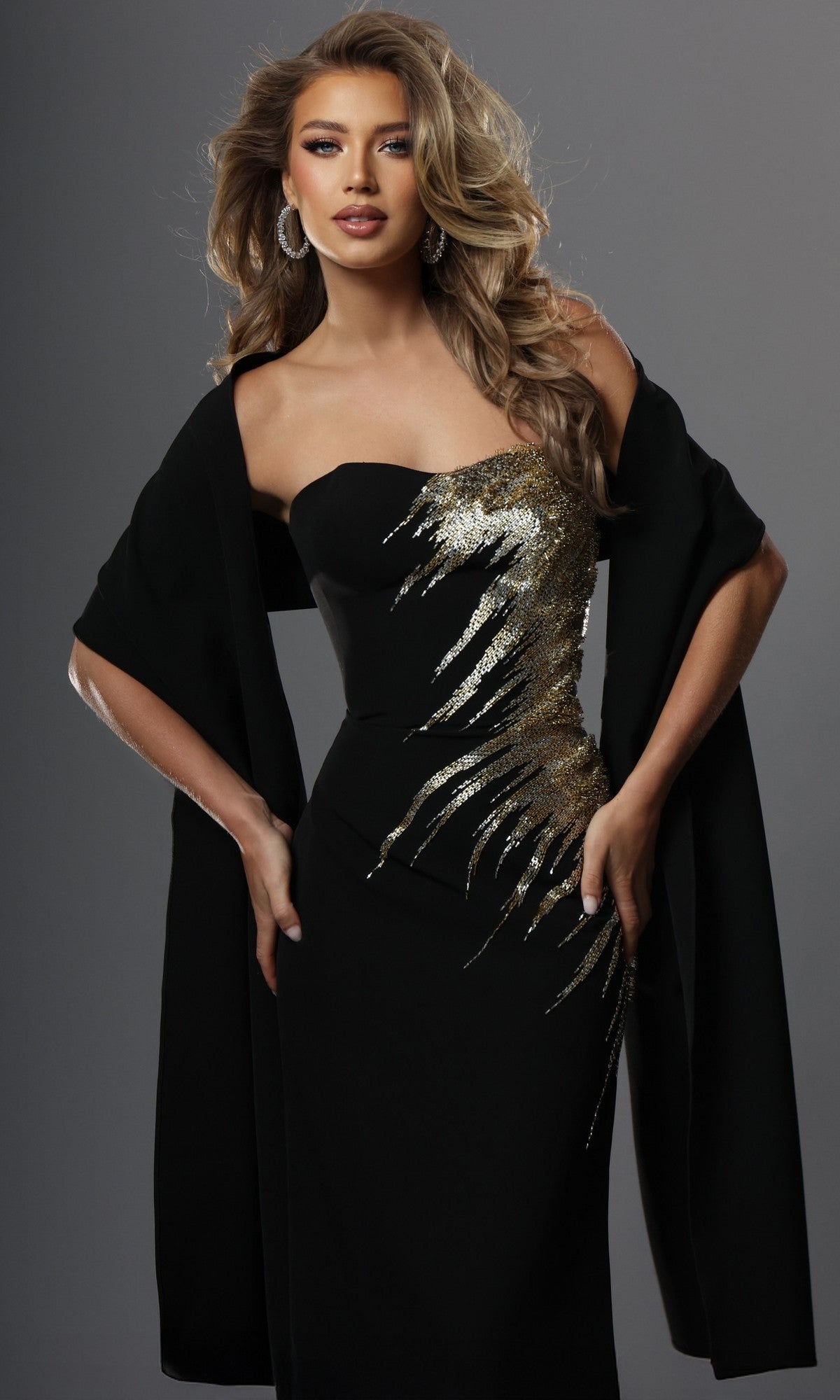 A woman poses against a neutral background wearing the Jovani 51900 long formal dress—a fitted sheath silhouette black strapless gown featuring gold sequin embroidery accents—styled with a matching black shawl draped over her arms.