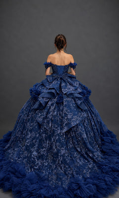 A woman stands facing away, wearing the Quinceanera Dress 5192 by Dancing Queen—a voluminous blue ball gown adorned with lace details, ruffles, and off-the-shoulder sleeves against a plain dark background. The shimmering beaded corset bodice adds an elegant touch to the timeless silhouette.
