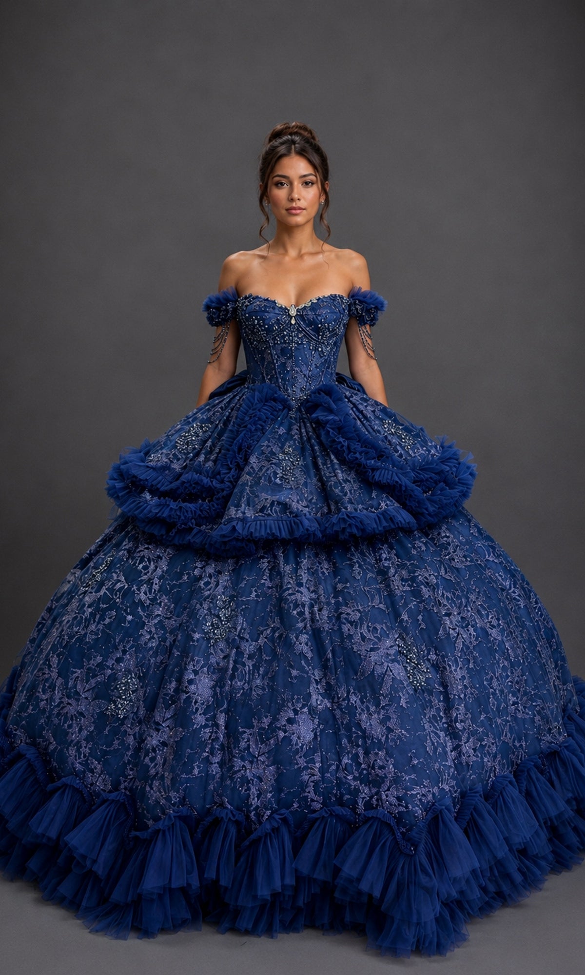 A woman wearing the Quinceanera Dress 5192 by Dancing Queen in navy, an ornate off-the-shoulder ball gown adorned with lace details and tulle accents, featuring a beaded corset bodice and a detachable layered skirt, stands against a plain dark background.