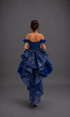 A woman stands with her back to the camera, wearing the Quinceanera Dress 5192 by Dancing Queen—a stunning blue ball gown featuring an off-shoulder layered design with intricate lace and tulle details, complemented by a detachable layered skirt, set against a plain gray background.