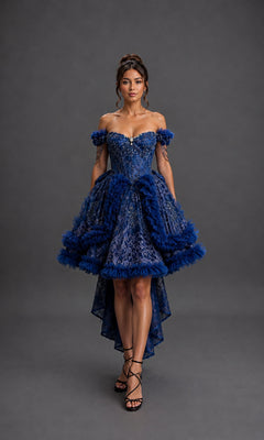 A woman stands facing forward against a gray studio backdrop wearing the Quinceanera Dress 5192 by Dancing Queen, an off-shoulder high-low blue lace dress featuring ruffles and a beaded corset bodice.